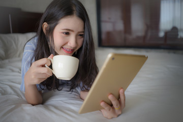 Happy Woman lying on bed using laptop drinking coffee. Young Asian girl watching movie on tablet.