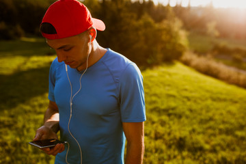 Young athlete male texting messages via his smart phone using free wireless in the park. Attractive runner listening to music during morning jog and run outdoor.