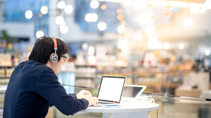 Young Asian business man listening to music by headphones while working with laptop computer in co working space. freelance or digital nomad lifestyle in urban workspace concepts - obrazy, fototapety, plakaty
