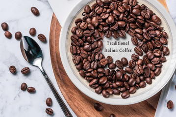 Traditional Italian Coffee beans are scattered around a white saucer and wooden chopping board, with stainless steel spoon.
