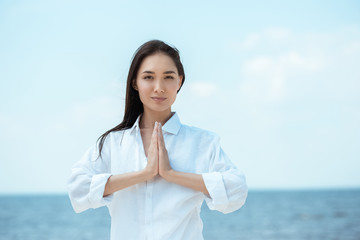 focused asian woman doing namaste mudra gesture in front of sea - obrazy, fototapety, plakaty
