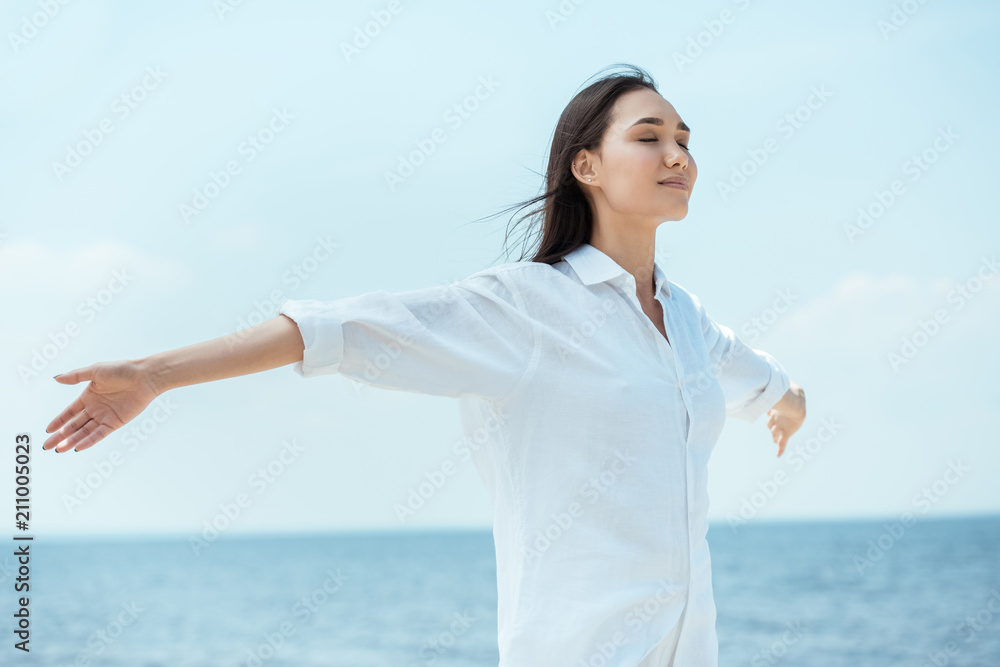 Wall mural young asian woman with closed eyes standing with arms outstretched by sea