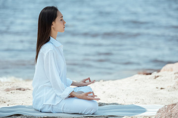 Side view of focused woman in ardha padmasana (half lotus pose) on yoga mat by sea