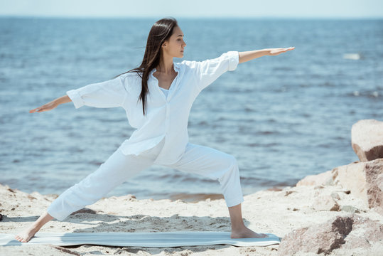 concentrated young asian woman practicing yoga in virabhadrasana (warrior II pose) on mat in front of sea