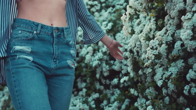 Low angle shot of a hot fit woman in trendy high-waist jeans walking down the garden of blooming flowers, touching the blossom. Beautiful landscape, active lifestyle, healthy living. Female portrait