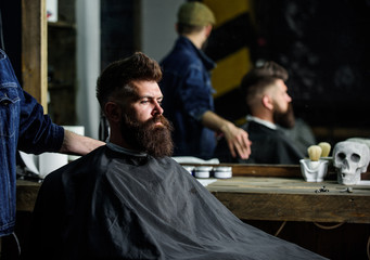 Man with beard covered with black cape sits in hairdressers chair, mirror background. Man with...