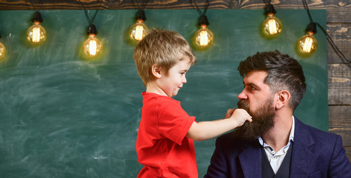 Teacher with beard, father and little son having fun in classroom, chalkboard on background. Child cheerful play with beard of teacher. Fatherhood concept. Dad with beard spend time with son.
