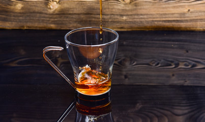 Cup pouring with water or tea with splashes on dark wooden background. Brewing tea concept. Glass with tea pouring with liquid with splashes and drops of water