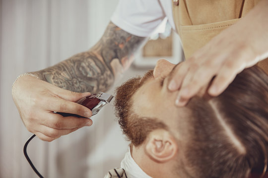 The barber's hand holds hair clipper while cutting the beard. Photo in vintage style