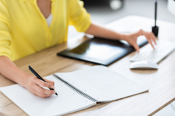 cropped image of female freelancer writing in textbook and working on computer at home office