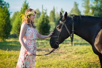 Poster Young woman walking with brown horse on meadow, wearing beautiful dress and midsummer wreath with field flowers © elinque