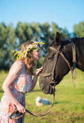 Obraz premium Young woman walking with brown horse on meadow, wearing beautiful dress and midsummer wreath with field flowers