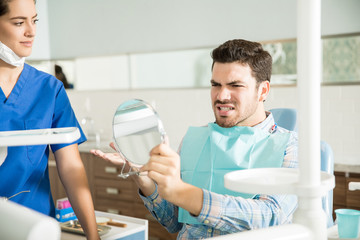 Man In Pain Looking At Mirror By Dentist In Clinic
