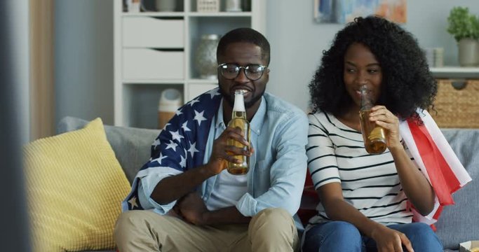 Portrait of the young smiled African American man and woman sitting on the sofa covered with an american flag and doing cheers gesture with beer. Indoor.