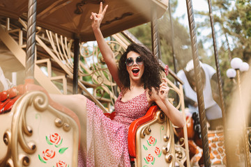 Young cheerful lady with dark curly hair in sunglasses and dress holding lolly pop candy in hand and showing two fingers gesture while riding on carousel in amusement park
