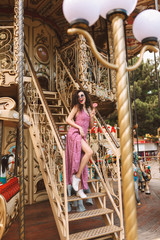 Pretty lady with dark curly hair in sunglasses and dress standing with lolly pop candy in hand and happily looking aside while spending time on beautiful carousel in amusement park