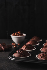 Chocolate muffins in bakeware with chocolate and nuts on the side on black concrete in front of a black wooden background with copy space.