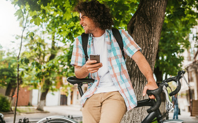 Attractive young hipster male with curly hair wearing shirt with backpack, sitting on the bike on the city street, browsing via Internet on smart phone, looking away, preparing for college. People