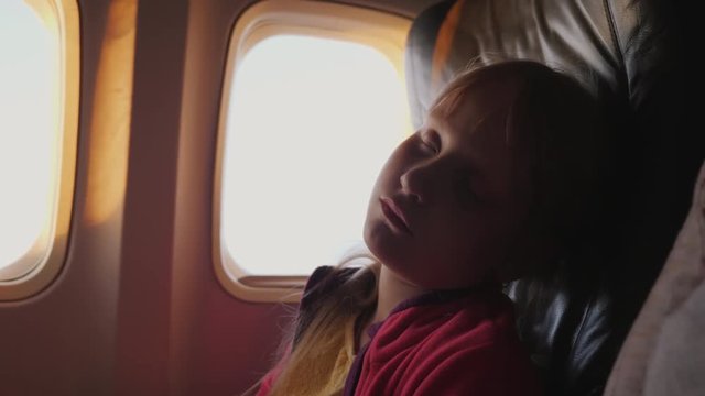 The little girl is sleeping in the cabin of the plane. During the dawn, the orange rays of the sun illuminate her face