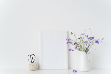 White portrait frame mockup with wild flowers in vase near white wall. Empty frame mock up for presentation design. Template framing for modern art.