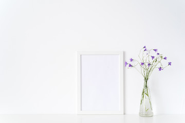 White portrait frame mockup with wild flowers in vase near white wall. Empty frame mock up for...
