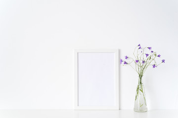 White portrait frame mockup with wild flowers in vase near white wall. Empty frame mock up for presentation design. Template framing for modern art.