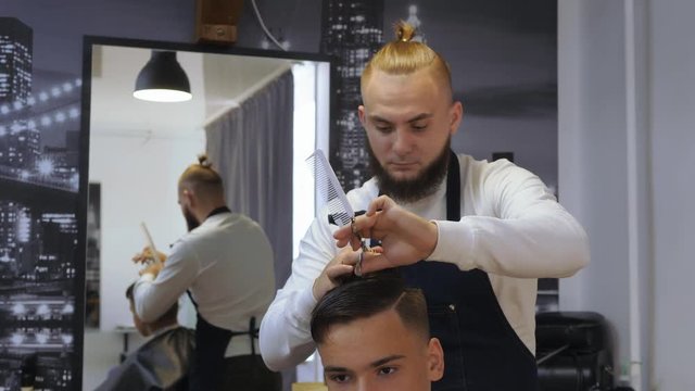 Hairdresser for men. Barbershop. A young guy gets a haircut and hair care service from a bearded man with a hair tied on his head. Cut hair head scissors with a comb. Funny hairdresser. Man