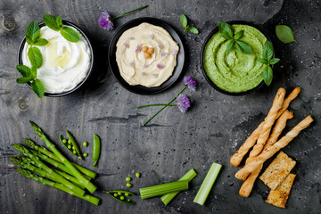 Green vegetables raw snack board with various dips. Yogurt sauce or labneh, hummus, herb hummus or pesto with crackers, grissini bread and fresh vegetables. Middle eastern meze snacks set