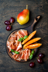 Jamon serrano or prosciutto with melon and fruits over rustic wooden background. Italian or spanish antipasti, appetizer board