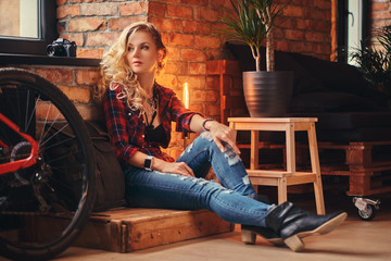 Obraz na płótnie Canvas Sensual blonde hipster girl with long curly hair dressed in a fleece shirt and jeans sitting on a wooden box, looking away, at a studio with loft interior.