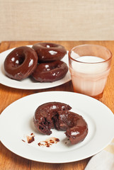 Top view, medium distance of a freshly baked, homemade chocolate donut broken in half on a white, round plate and a glass of milk, and fore more donuts in background 