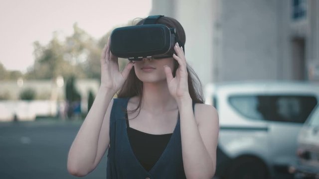 Portrait of attractive woman looking around using VR glasses on street. Girl getting expirience while wearing virtual reality headset at summer outdoor.