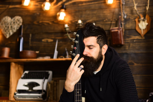 Connection through music. Man bearded musician enjoy evening with bass guitar, wooden background. Guy sits dreamy in cozy atmosphere. Man with beard hugs neck of electric guitar. United with music