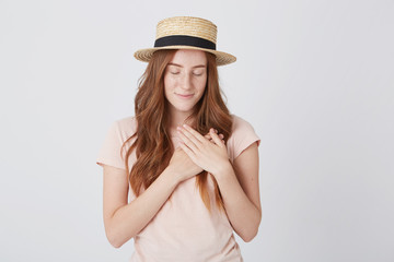 Smiling tender redhead young woman in straw hat and tshirt standing with eyes closed and hands on her heart isolated over white background