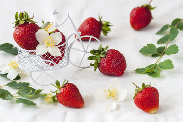 Miniature toy shopping cart, trolley with ripe strawberries, leaves, flowers on light background. Vegetarian concept, diet, detox, organic vitamins