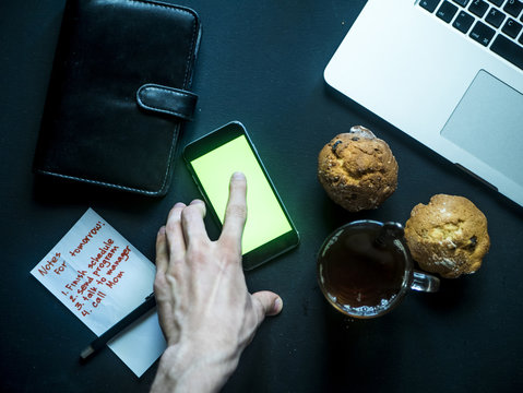 Top View Of Finger Scrolling Mobile Smartphone On Dark Table With Laptop And Note