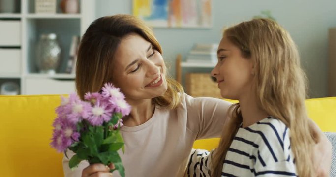 Close up of the pretty teenager girl bringing flowers to her attractive mother and hugging her on the sofa in the living room. Indoors.