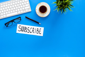 Email subscribe concept. Hand lettering subcribe on work desk with plant, glasses, computer keyboard, cup of coffee on blue background top view copy space