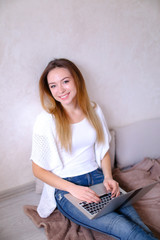 Young woman wearing white t shirt and jeans, typing by laptop and sitting on bed. Concept of...