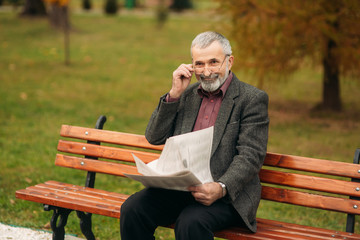 Fototapeta premium A nice grandfather with a beautiful beard in a gray jacket sits on a bench in the park and reads a newspaper. Loot at the photographer