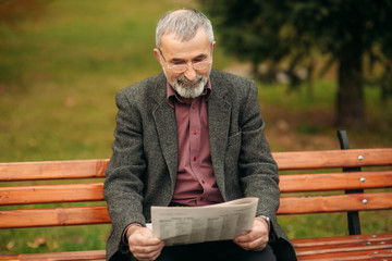 Fototapeta premium A nice grandfather with a beautiful beard in a gray jacket sits on a bench in the park and reads a newspaper