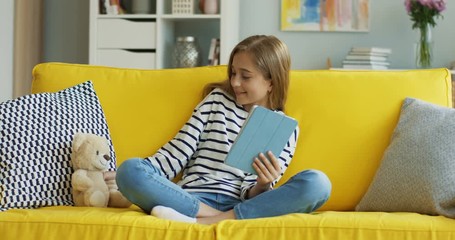 Cute teen blonde girl sitting on the yellow couch at home with a tablet computer in hands and fixing her teddy bear near her. Inside.