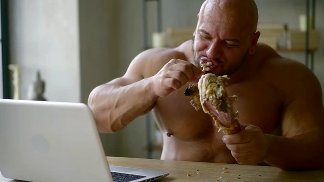 powerful sports bald man with a large physique eating a large piece of meat bird pulling his teeth from the bone while working at the laptop