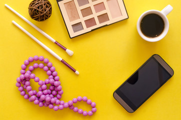 Color eyeshadow palette, mobile phone with blank screen, make up brush and cup of hot cocoa on yellow background.