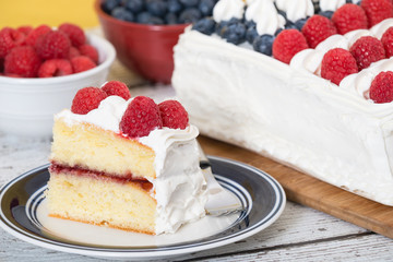 A slice of patriotic, red white and blue, American flag cake. The cake with fresh blueberries and raspberries in the background. 