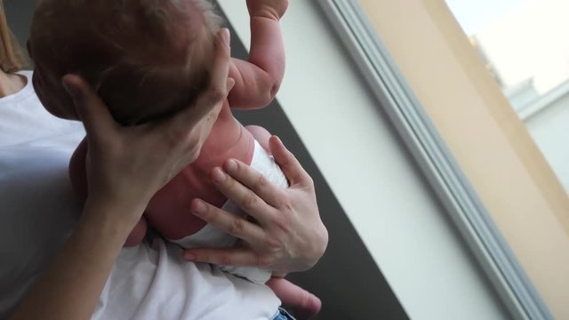 Young mother holds her first newborn baby son stroking and wiggling him on hands by a window