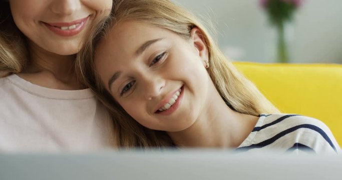 Close up of the cute smiled teenage girl leaning on her mother's shoulder while watching something on the laptop computer. Indoor.
