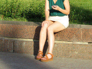 Woman in a white mini skirt sitting with a smartphone in her hand. Waiting girl, female naked legs, summer fashion, online dating, reading woman