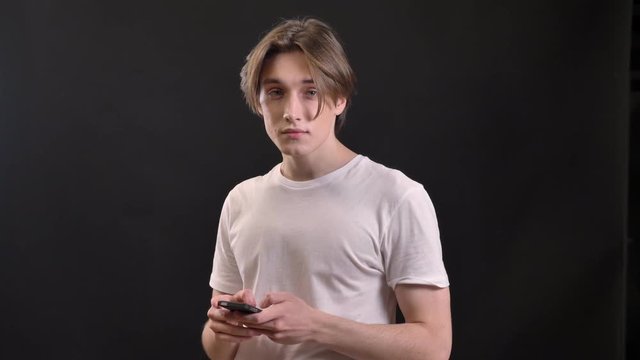 Young charming man in white shirt holding phone and looking at camera, isolated over black background
