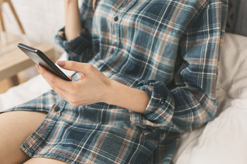 Beautiful girl is sitting on the white bed linen. Young woman in men's checkered shirt is searching in social network on white pillow. Morning with black smartphone in hands.